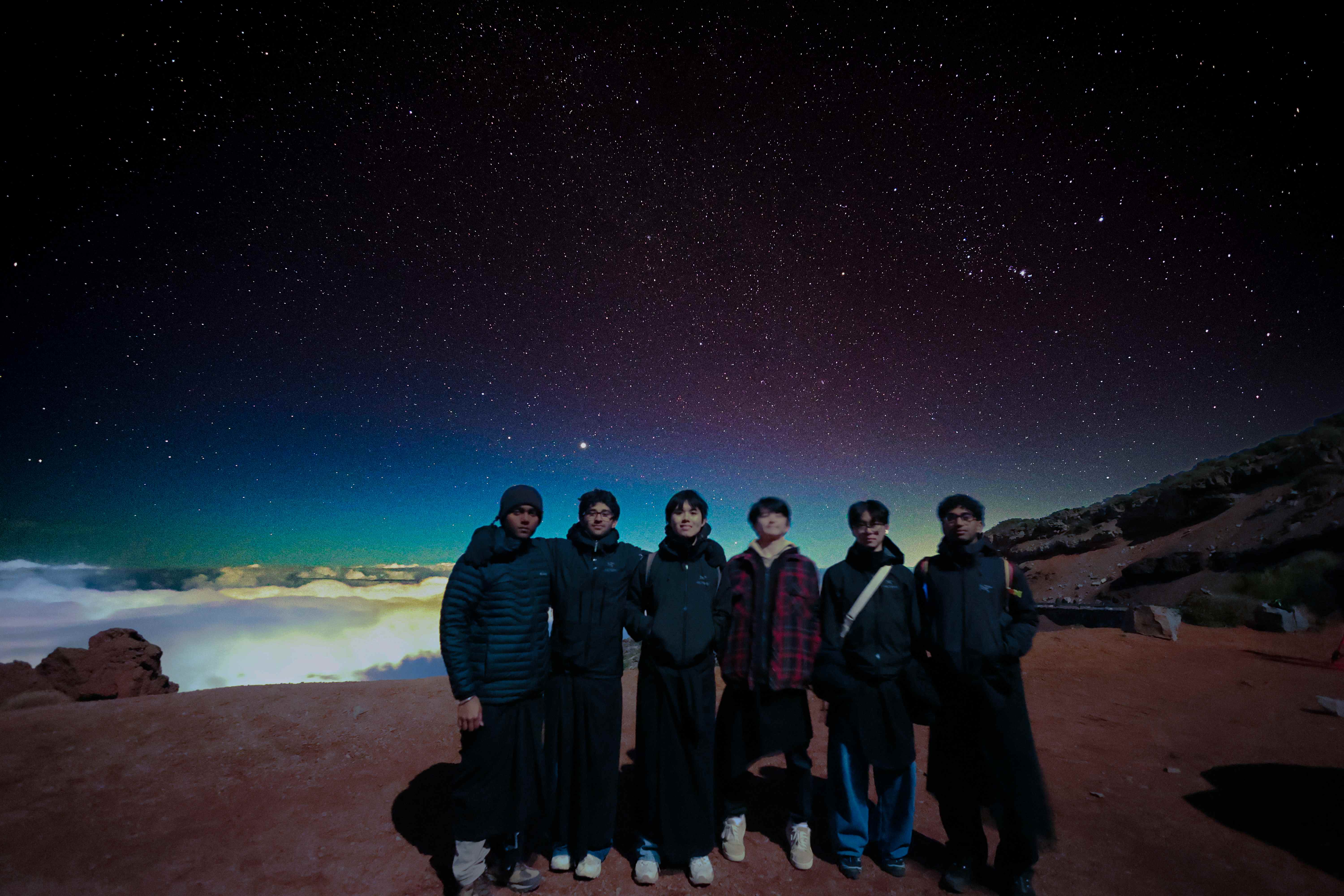 Group of six friends on a volcanic summit at night above the clouds, teal aurora glow on the horizon