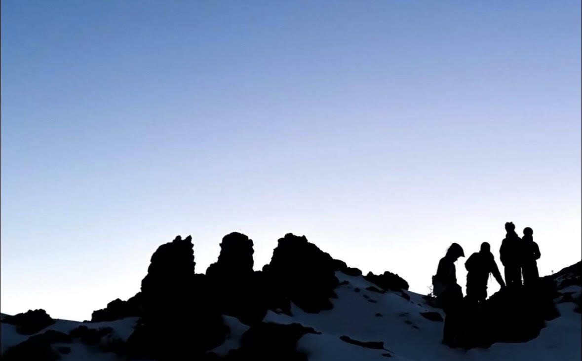 Silhouettes of lava rock formations and a group of people on a snowy ridge against a pale blue pre-dawn sky